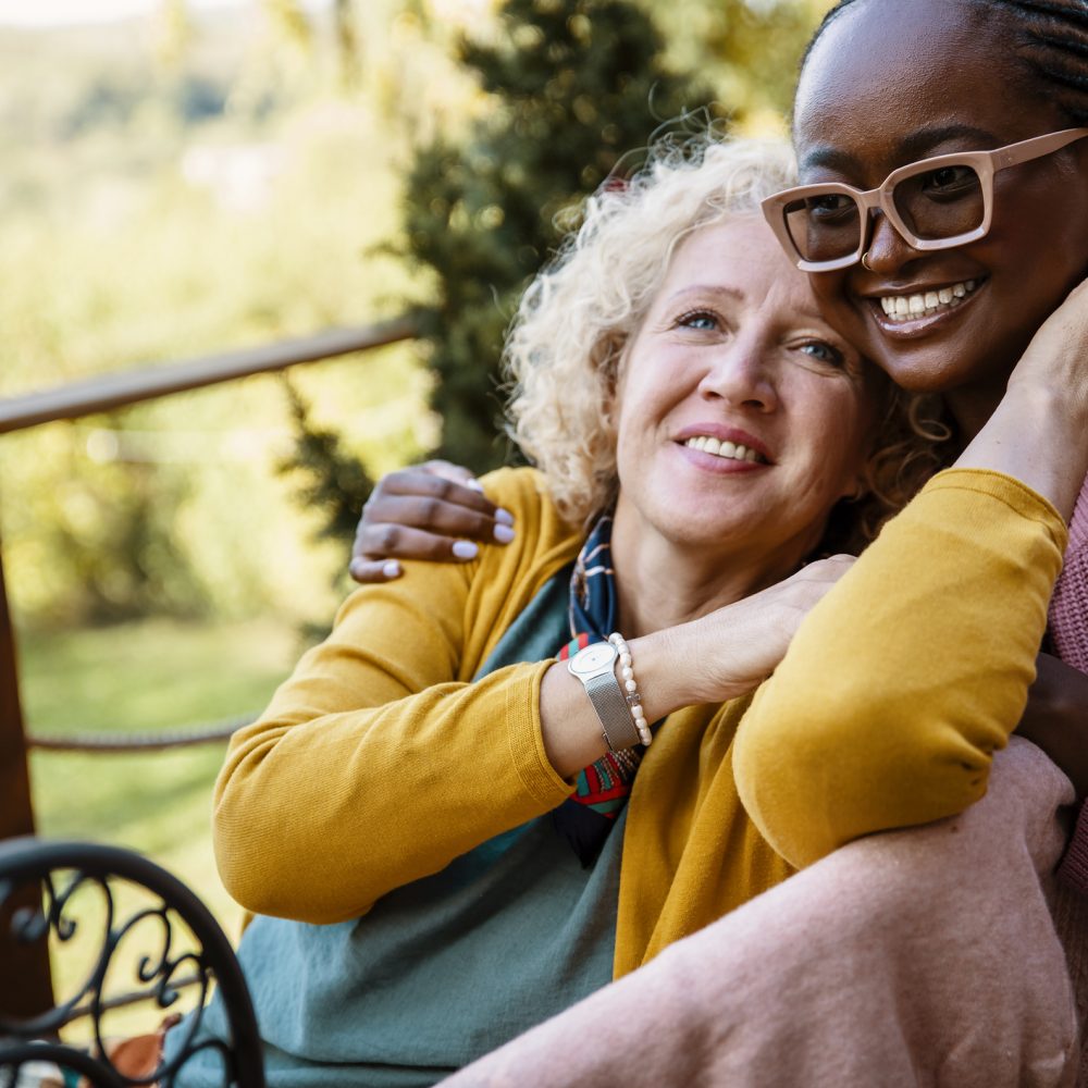 Mixed race female family relaxing in rehabilitation center in nature. Young African American Woman take care of disabled older Caucasian mother on a porch of nursing home. Copy space